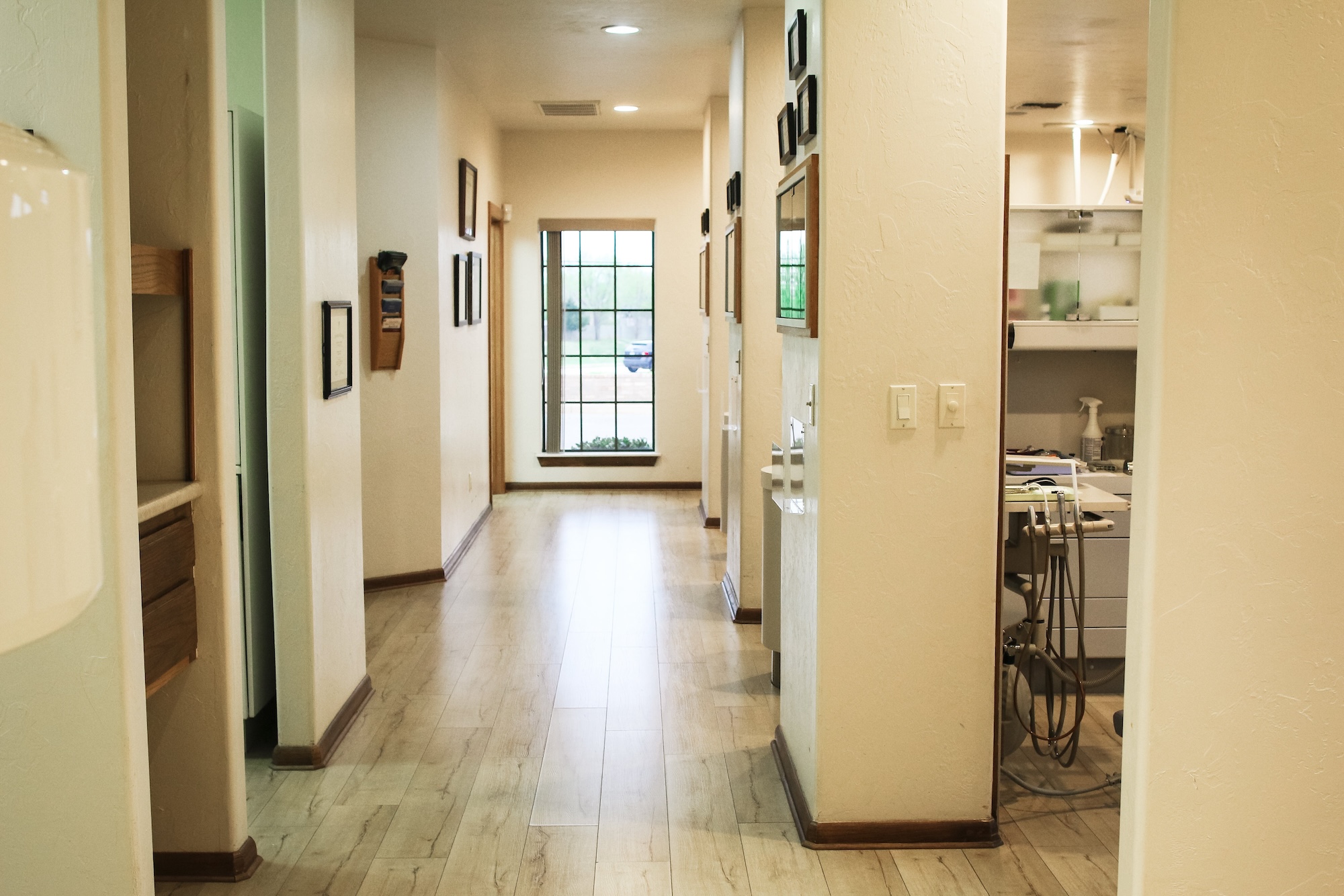 The image shows an interior hallway with shiny wooden floors, walls adorned with framed pictures, and a large window allowing natural light into the space.