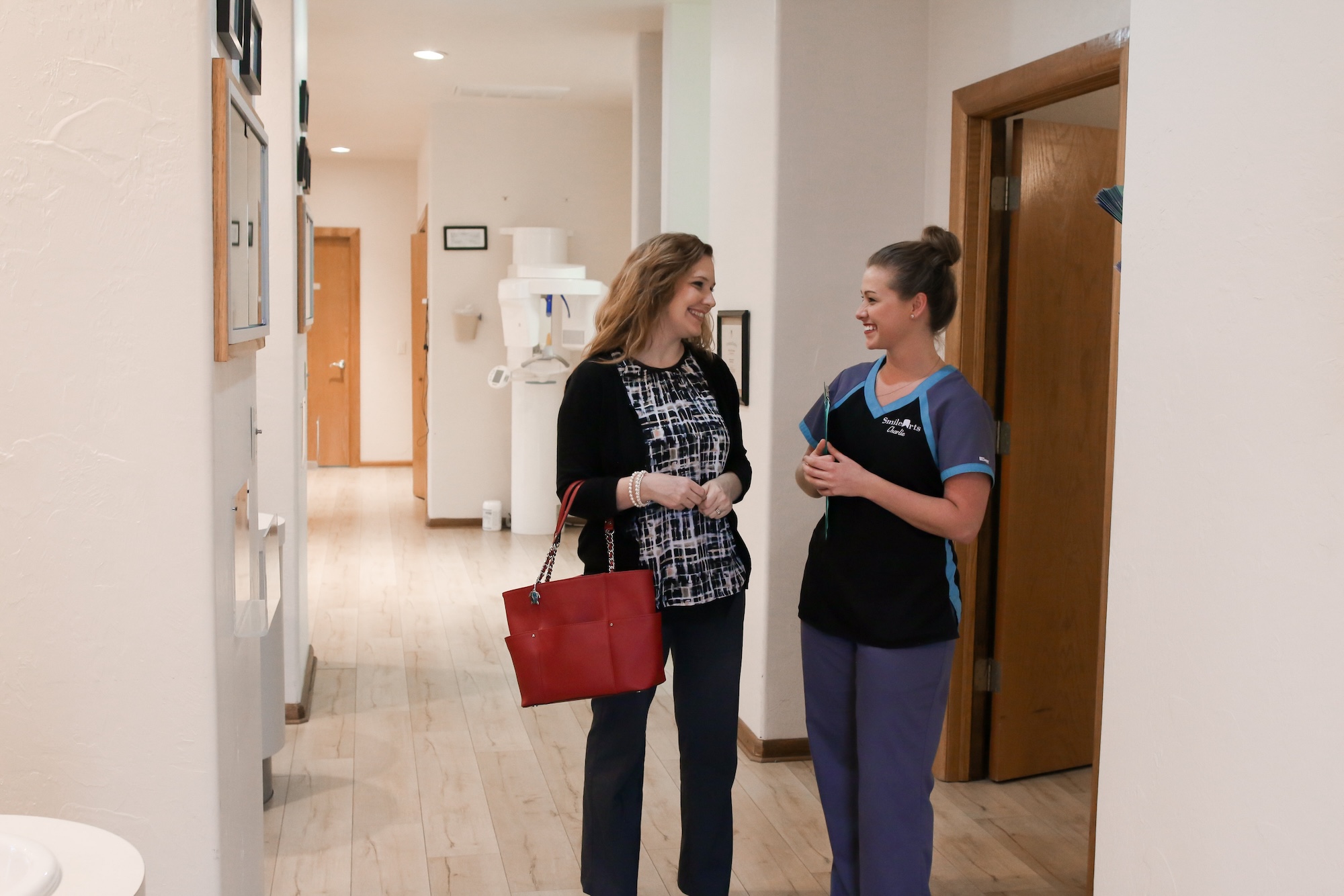 A woman and a man are standing in an indoor hallway within a building, with the woman wearing a black shirt and jeans, and the man dressed in a white shirt and blue pants, both smiling at each other.