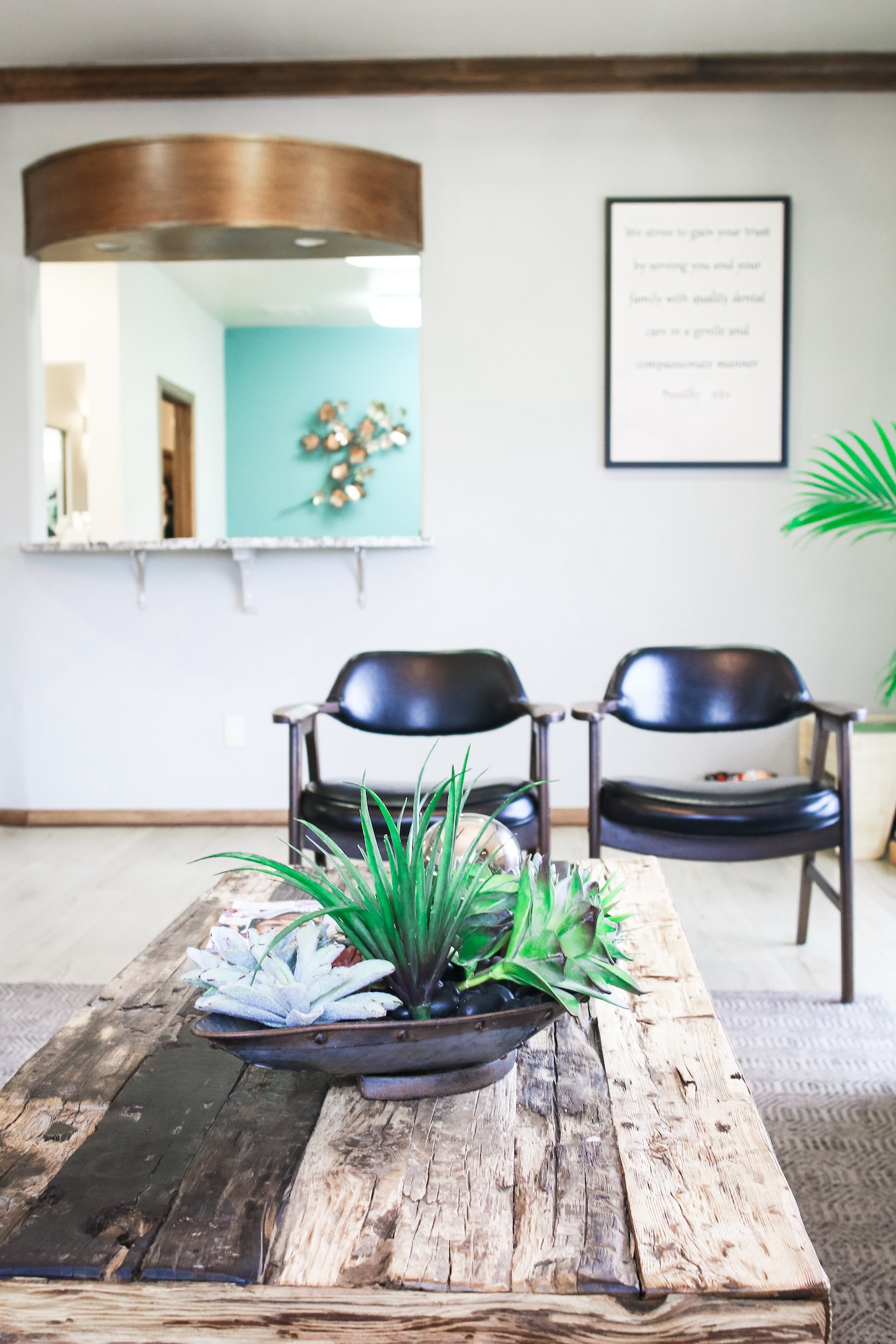 The image depicts an interior space with a wooden table at its center, surrounded by chairs, featuring a modern design aesthetic with exposed brick walls, plants, and a framed piece of artwork on the wall.
