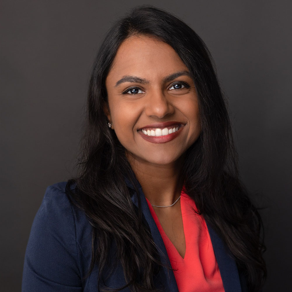 A woman with a smile, wearing a blue blazer over a red top, poses for a portrait against a dark background.