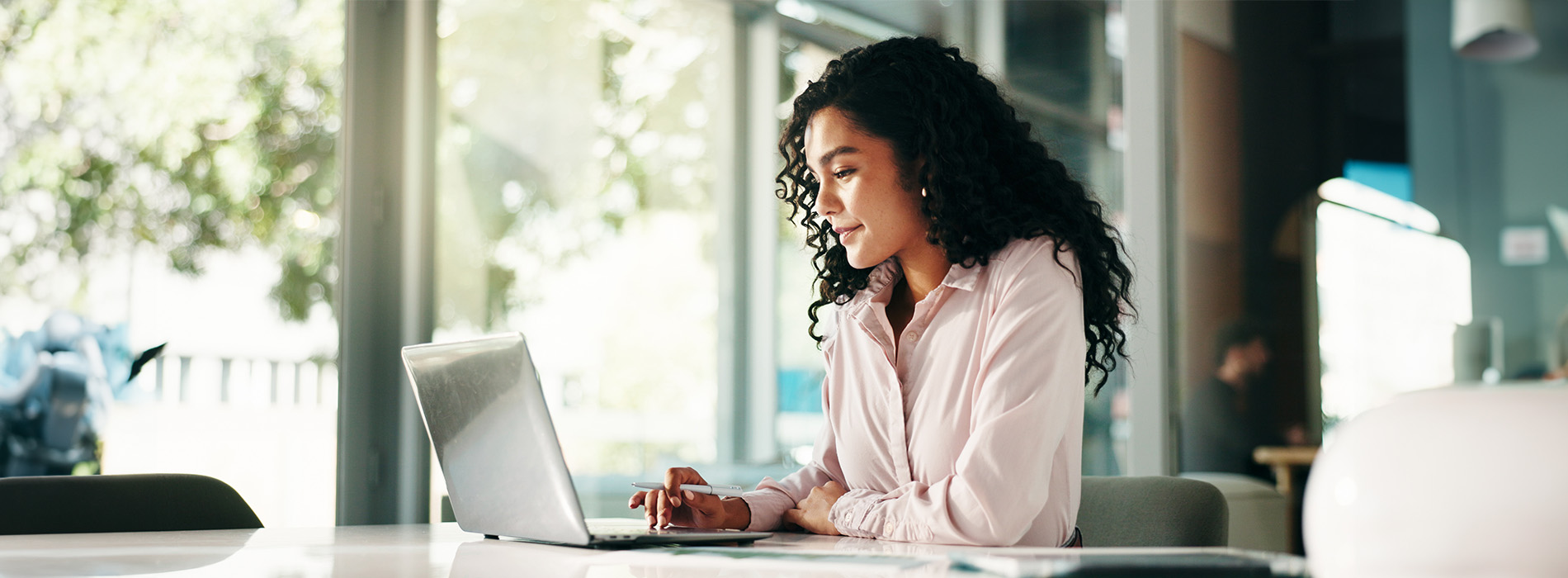 The image features a woman seated at a desk with a laptop, working in an indoor setting with natural light coming through a window.
