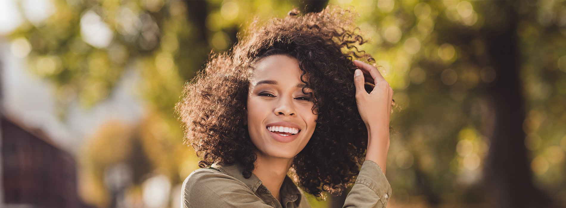The image shows a young woman with curly hair smiling at the camera while standing outdoors during daylight. She has her hand on her head and appears to be looking upwards.