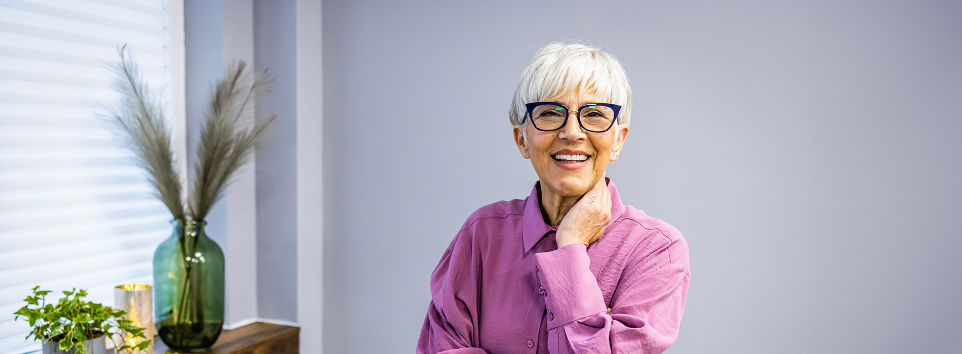 An elderly woman wearing glasses and a purple shirt stands in front of a window with plants, smiling at the camera.