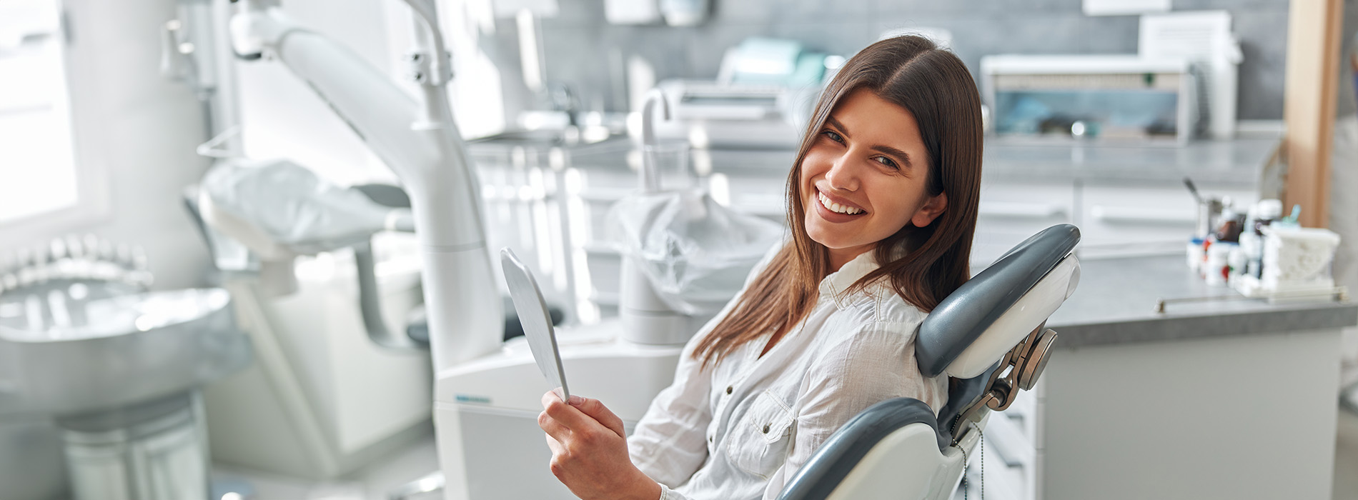 A woman seated in a dental chair with a smile, holding a tablet in her hand, surrounded by dental equipment and a dental office setting.