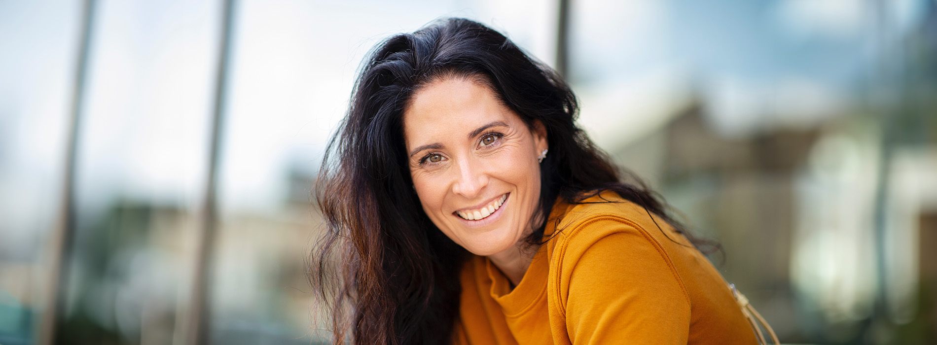 The image shows a smiling woman with long hair, wearing a yellow top, against an indoor background with natural light.