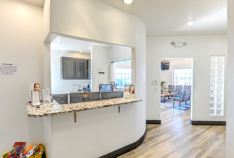 The image displays an interior view of a spacious waiting area within a building, featuring a reception desk with a countertop, a sign above the counter, a large window allowing natural light into the space, and a modern design aesthetic.