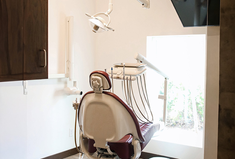 The image shows a dental office interior with a chair and dental equipment in the foreground, and a window in the background letting in natural light.