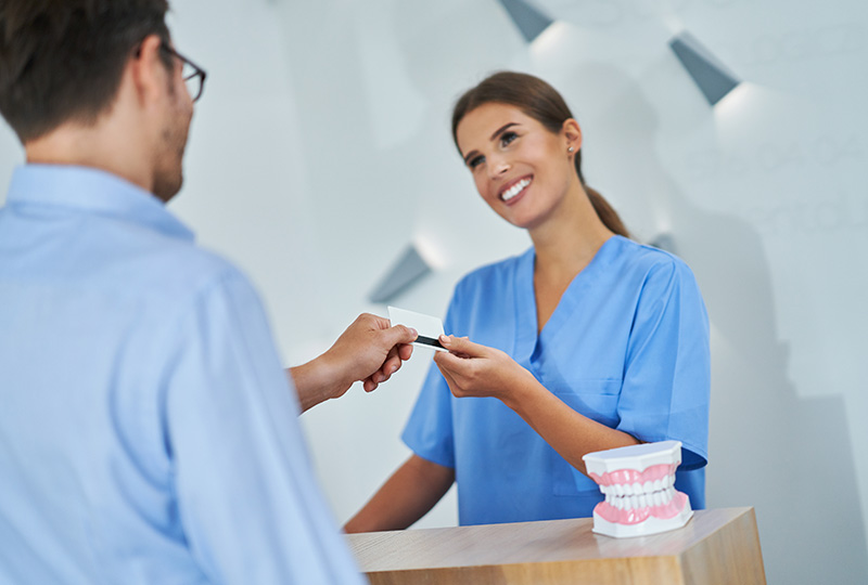 A woman in blue scrubs hands over a card to a man at a desk with a toothpaste tube on it, both smiling.
