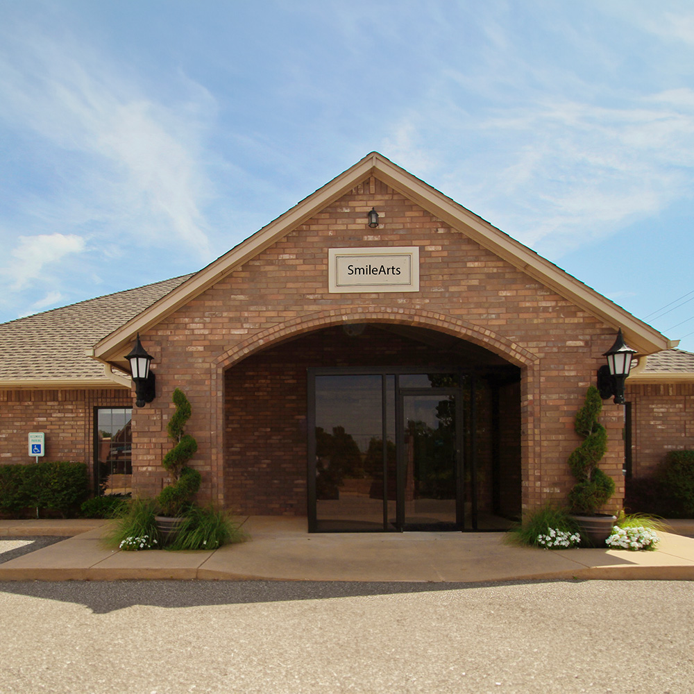 The image shows a brick building with a sign that reads  Smile Art  on its front, featuring a white frame, a black lamp post, and a paved driveway with a clear sky in the background.