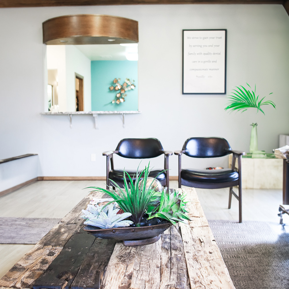 The image shows a modern interior space with a wooden table, chairs, potted plants, and decorative items such as vases and books. A wall-mounted sign with text is visible above the table.