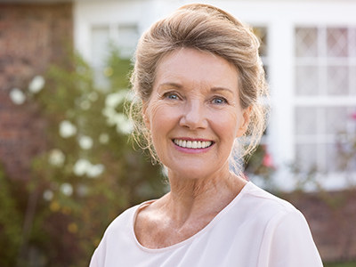 The image shows a woman standing outdoors, smiling at the camera, with her hair styled up, wearing a white top, and posing in front of a house with a brick wall and greenery.