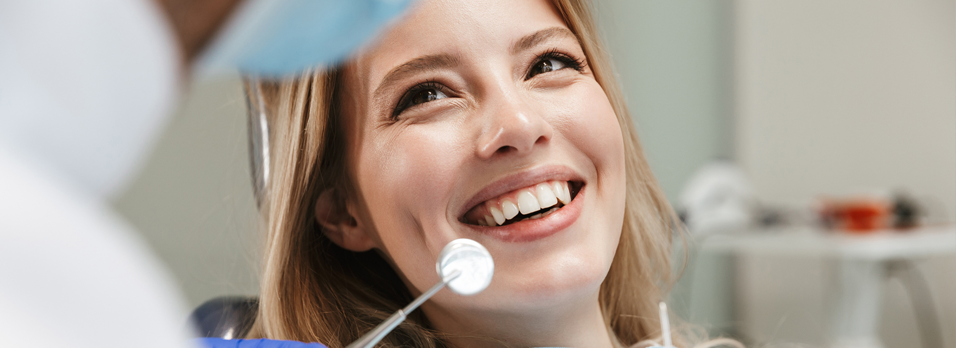 The image shows a young woman with a big smile looking at a camera, sitting in a dental chair with a dentist behind her, who appears to be examining her teeth.