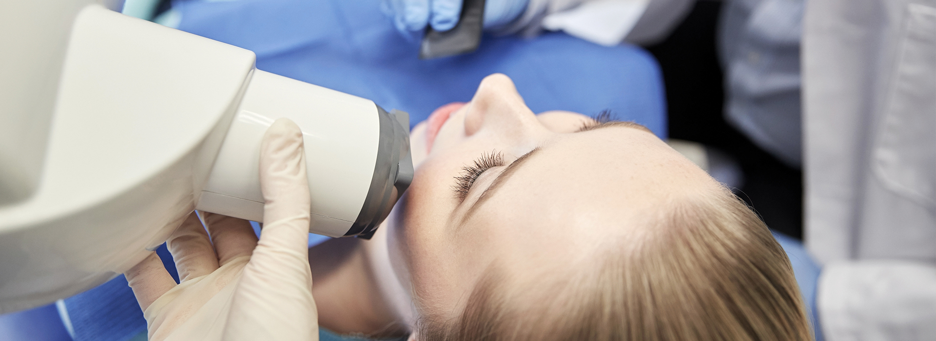 A person s head being examined under a microscope by a medical professional wearing gloves and a blue surgical mask, with a focus on the examination process.