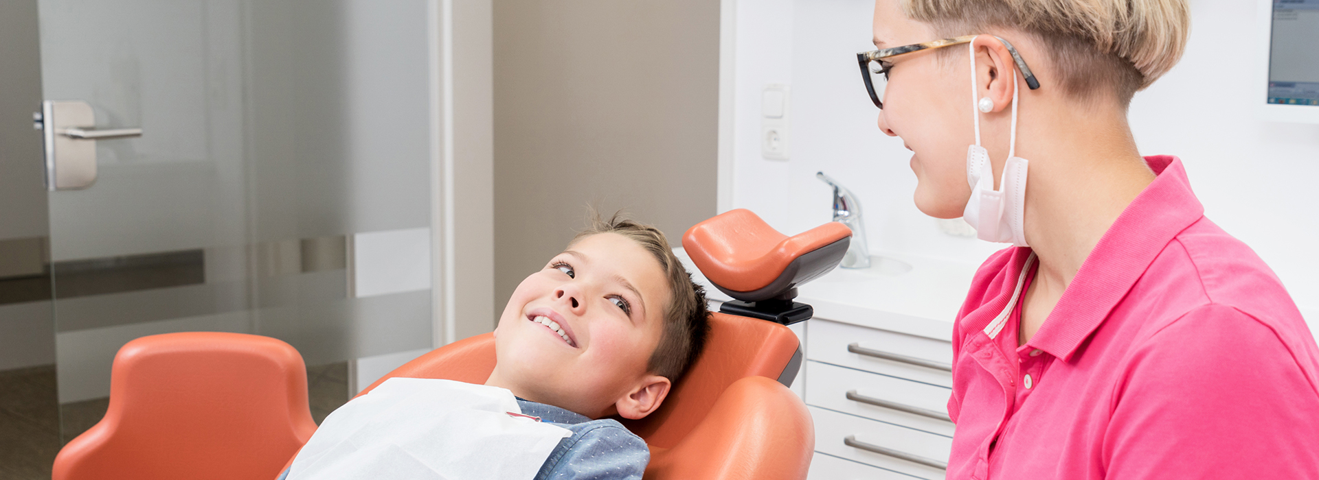 The image shows a dental office setting with a woman sitting in a dental chair receiving dental care from a dentist, who is standing behind her.