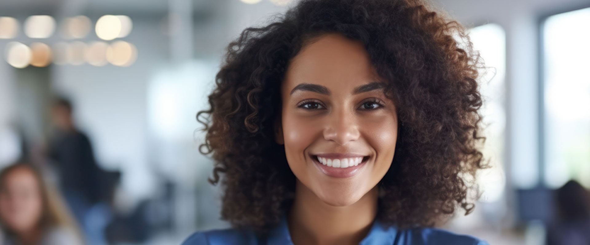 The image features a smiling woman with curly hair, wearing a blue top, standing in an office environment, looking directly at the camera.