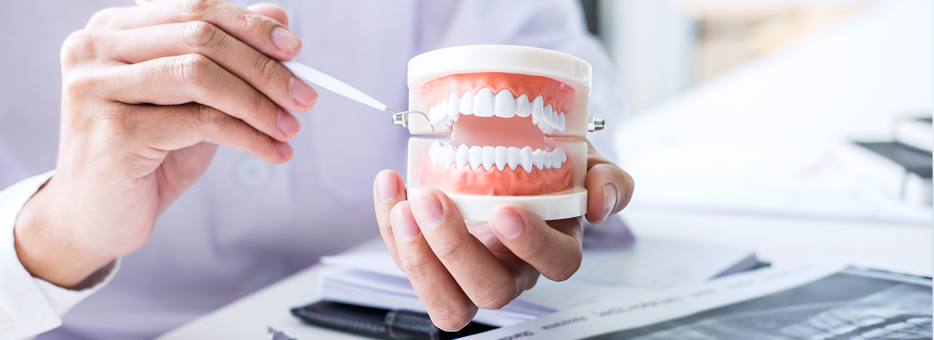 A person s hand holding a toothbrush over a cup containing a toothpaste-filled mouthguard, with another hand holding a pen and paper behind the cup, against a background of a blurred office environment.