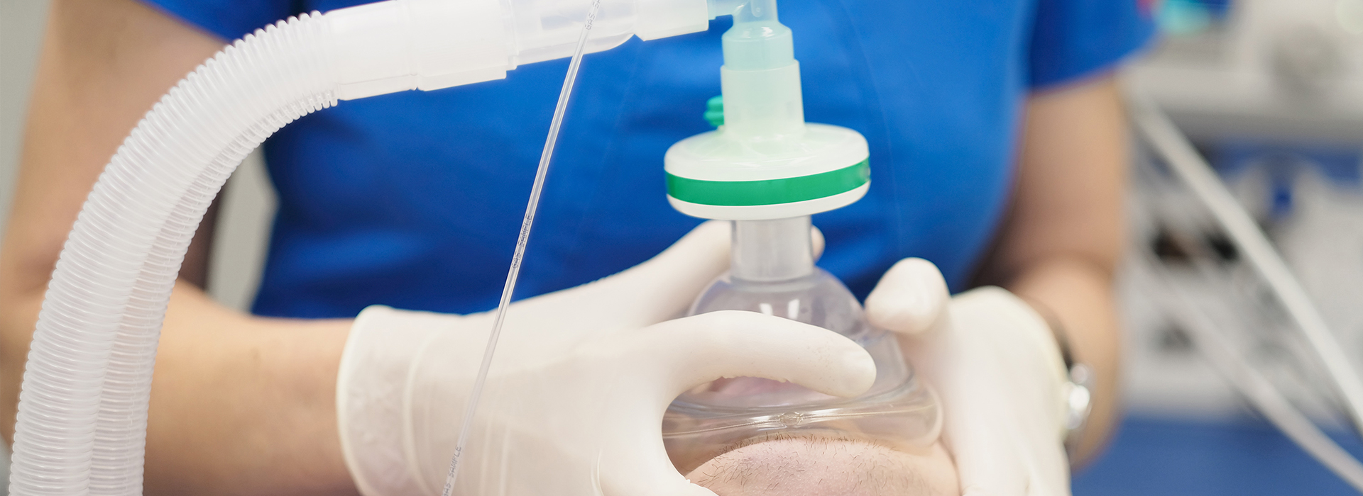 A medical professional wearing gloves and a face mask administers medication through an IV tube while standing at a hospital workstation.