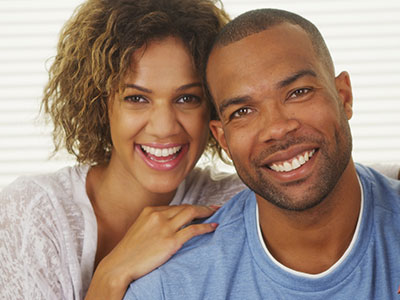 The image shows a man and woman posing together with smiles  the man is wearing a dark-colored shirt and has short hair, while the woman has curly hair and is wearing a light-colored top.