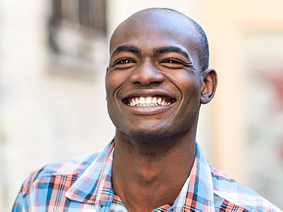 The image shows a young man with a big smile, wearing a plaid shirt, standing outdoors.