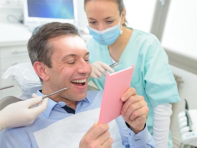 A man sitting in a dental chair with a pink cardboard prop, smiling at the camera while being attended by two dental professionals.