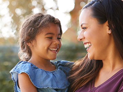 The image shows a woman with a child, both smiling at the camera, against a blurred background.