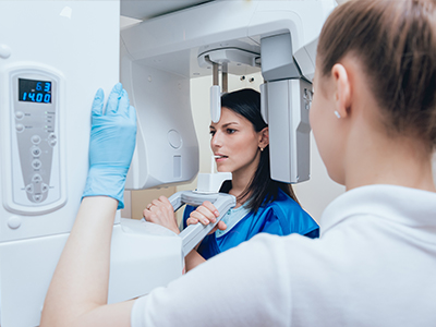 A woman stands next to a large CT scan machine with a digital display showing  140  on its screen, while another woman, presumably an employee, looks at her with interest.