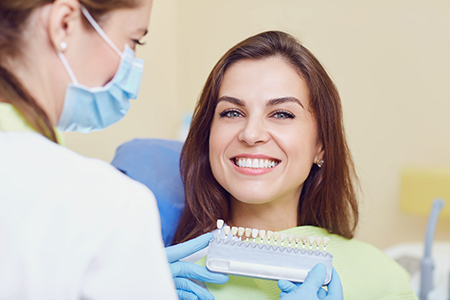 A woman is smiling at the camera while sitting in a dental chair, receiving dental care from a professional who is holding a device near her mouth.