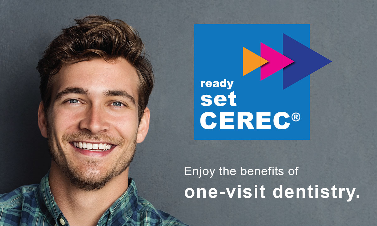 A man with a beard smiles at the camera, positioned in front of a blue sign with white text that reads  READY SET CEREC  and below it,  enjoy the benefits of revisit dentistry.