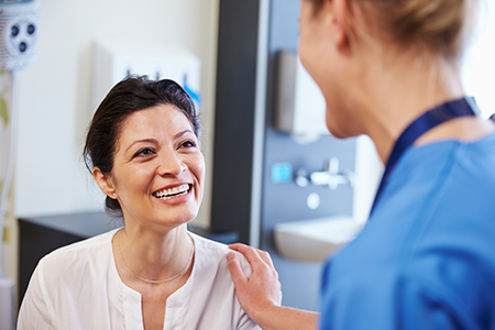 The image shows two individuals in a medical setting  a woman with a smile, wearing a white top, seated next to a person who appears to be a healthcare professional, possibly a nurse or doctor, wearing a blue scrubs and smiling at her.