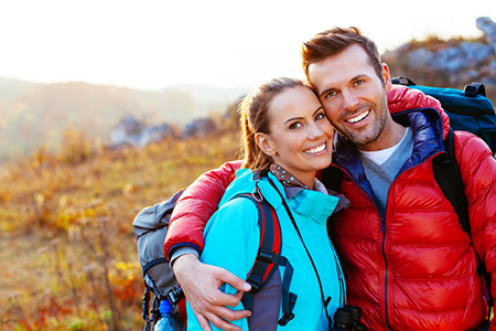 The image shows a man and a woman posing for a photo with backpacks, smiling and embracing each other against a backdrop of natural scenery during daylight.