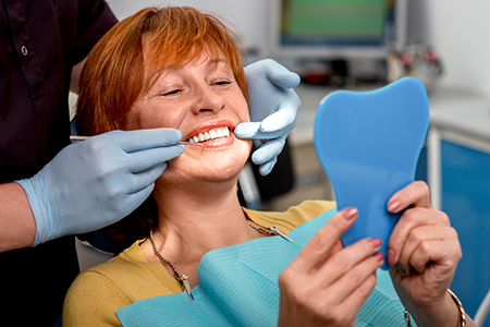 A woman sitting in a dental chair with a blue mouthguard on her teeth, smiling at the camera while a dentist adjusts her smile.