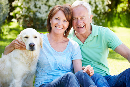 A man and woman are sitting on grass with a golden retriever between them, smiling at the camera.