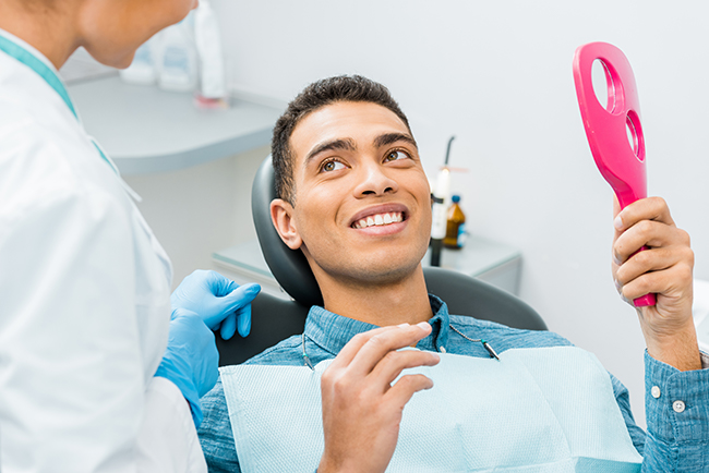 The image depicts a dental professional sitting in a chair with a patient s head, smiling at the camera while holding a pink object that resembles a toothbrush.