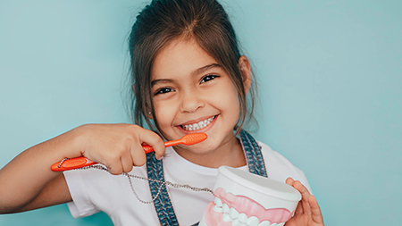 The image shows a young girl brushing her teeth with an oversized toothbrush while holding a small toy toothbrush, smiling broadly, and looking directly at the camera.