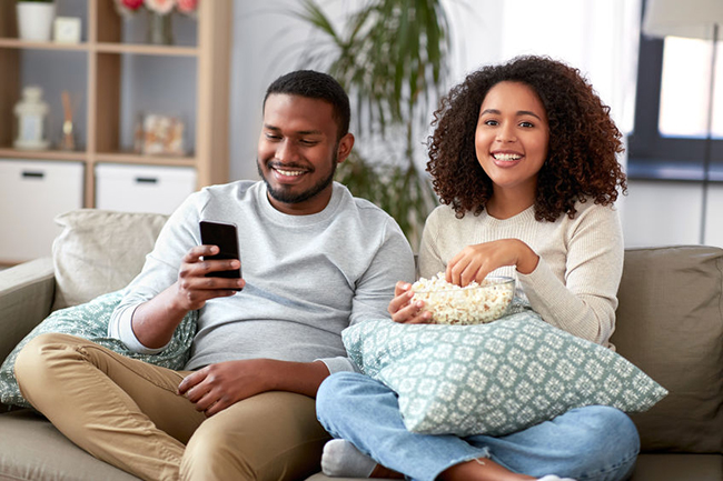 A man and woman sitting on a couch, smiling at each other while holding snacks, with a cell phone between them displaying a photo of another couple.