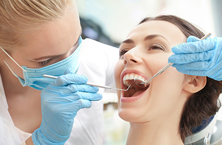 The image shows a dental hygienist performing a teeth cleaning procedure on a patient s mouth with an electric toothbrush, wearing protective gloves and using a dental mirror to inspect the teeth.