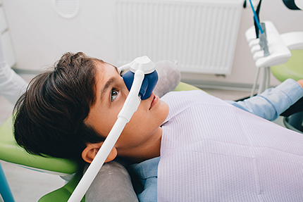 The image shows a young person sitting in a dental chair with a dental mirror over their eyes, wearing a mouthguard, and looking upwards towards the camera.