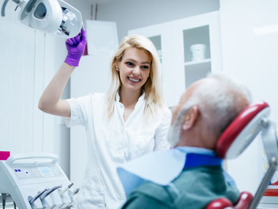 A woman in a white coat stands next to an older man seated in a dental chair, holding a dental mirror over his face.