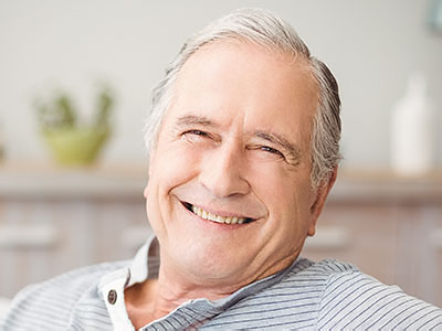 A smiling older man with gray hair, wearing a blue shirt, leaning back on his couch with his eyes closed.
