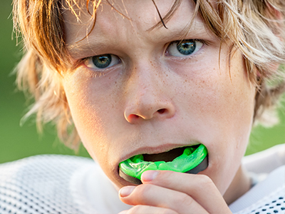 A young boy with blonde hair holds a green toothbrush in his mouth while posing for a photo, with the background showing a football field.