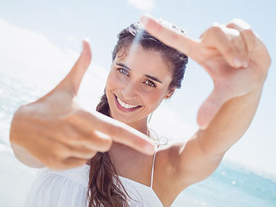 A young woman with long hair smiles while holding up her hand towards the camera, creating a frame with her fingers.