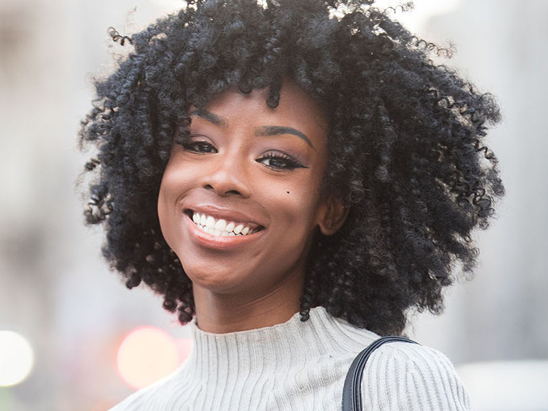 The image features a woman with curly hair smiling at the camera, wearing a dark top and a light-colored scarf around her neck.